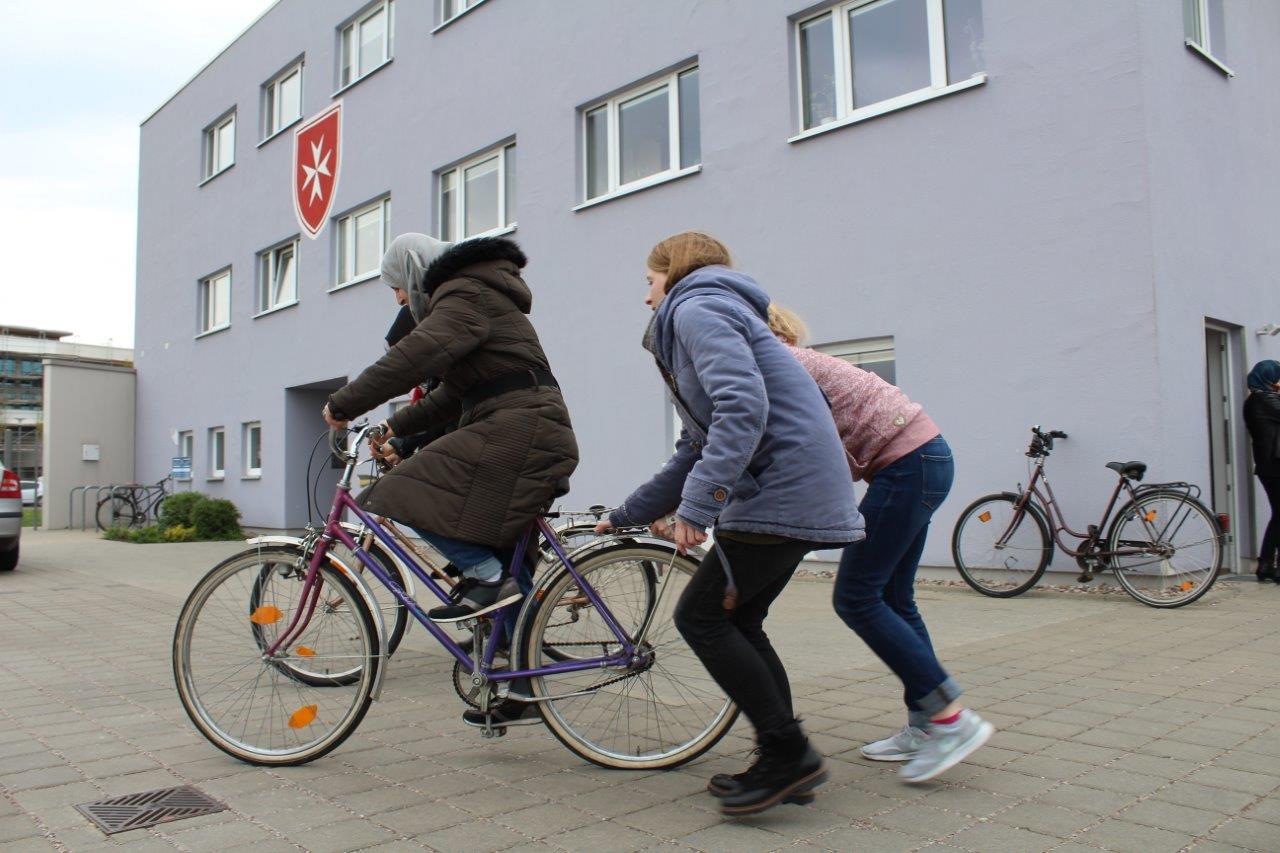 Zwei Frauen schieben eine andere Frau an, die auf einem Fahrrad sitzt.